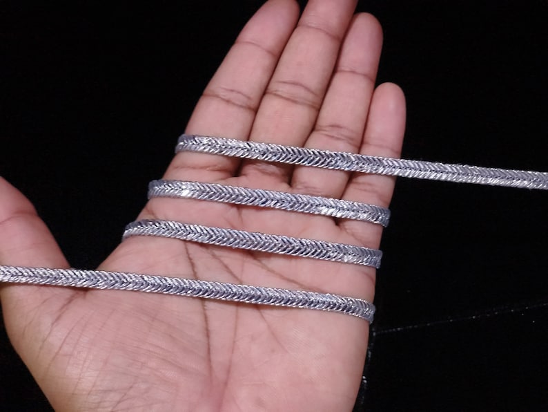 May include: Silver trim with a herringbone pattern. The trim is shown in a hand, with multiple strands of the trim.
