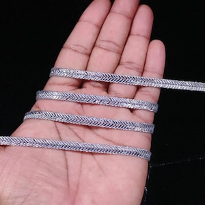 May include: Silver metallic trim with a chevron pattern. The trim is shown in a hand, wrapped around itself several times.