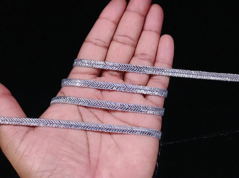 May include: Silver metallic trim with a chevron pattern. The trim is shown in a hand, wrapped around itself several times.