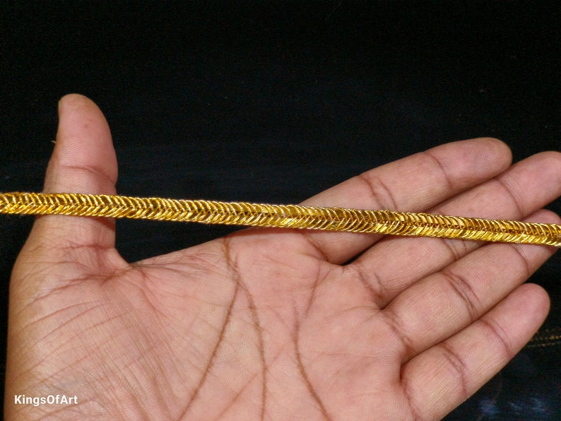 May include: Gold trim with a zig-zag pattern. The trim is shown on a hand.