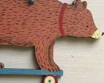 May include: A brown wooden bear ornament with a red collar and painted fur texture, mounted on a blue skateboard with four wheels featuring red rims and cream centres. The bear is facing right and has a cream-coloured snout with black dots.