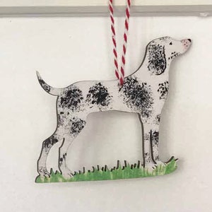 May include: A white and black spotted dog ornament with a red and white string hanging from the top. The dog is standing on a green patch of grass.
