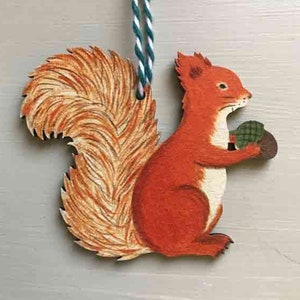 May include: A wooden squirrel ornament with a red body, a bushy tail, and a green acorn in its paws. The ornament is hanging from a white and green string.