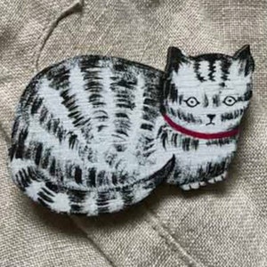 May include: A black and white painted wooden cat brooch with a red collar. The cat is lying down with its head turned to the side. The brooch is on a light brown fabric background.