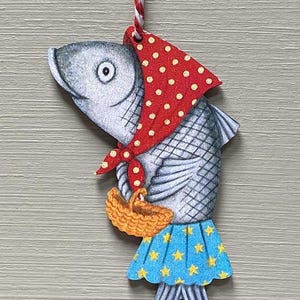 May include: A charming wooden fish decoration, wearing a red spotted scarf and a blue skirt with yellow stars. The fish holds a small basket and hangs from a red and white string, ideal for festive displays.