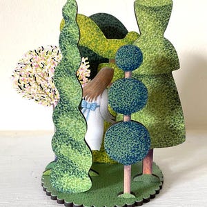 May include: A wooden figurine of a girl standing in a woodland setting. The girl is wearing a white dress with a blue bow. The woodland is made up of green trees and bushes. The figurine is painted in a realistic style.