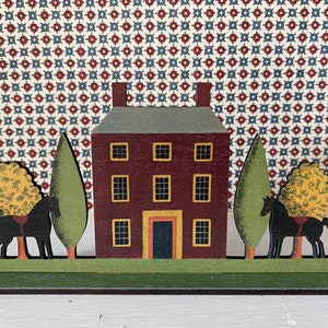 May include: A wooden folk art style house with a red brick facade and a green roof. Two black horses stand on either side of the house, each with a yellow tree behind them. The house has four windows and a door with a yellow trim. The base of the house is green.