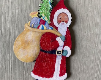 May include: Wooden Christmas ornament depicting Santa Claus carrying a sack of toys. Santa is dressed in a red coat with white trim, a black belt, and blue boots. The ornament includes a green and white string for hanging.