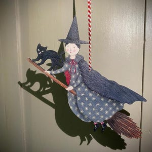 May include: A wooden Halloween decoration featuring a witch riding a broom with a black cat. The witch wears a blue dress with white stars, a gray cape, and a pointed hat. The decoration hangs from a red and white string.