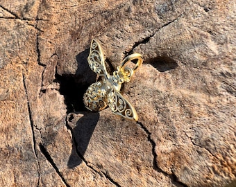 May include: A gold-colored bee charm with intricate details, featuring a detailed body and wings. The charm is displayed on a rustic wooden surface.