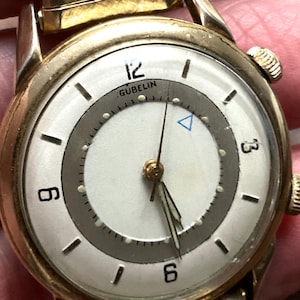 May include: A vintage gold-toned wristwatch with a white face and a gray inner ring. The watch face features black hour markers, the brand name "GUBELIN", and a blue triangle. The watch has a gold-toned band and a textured winding crown.