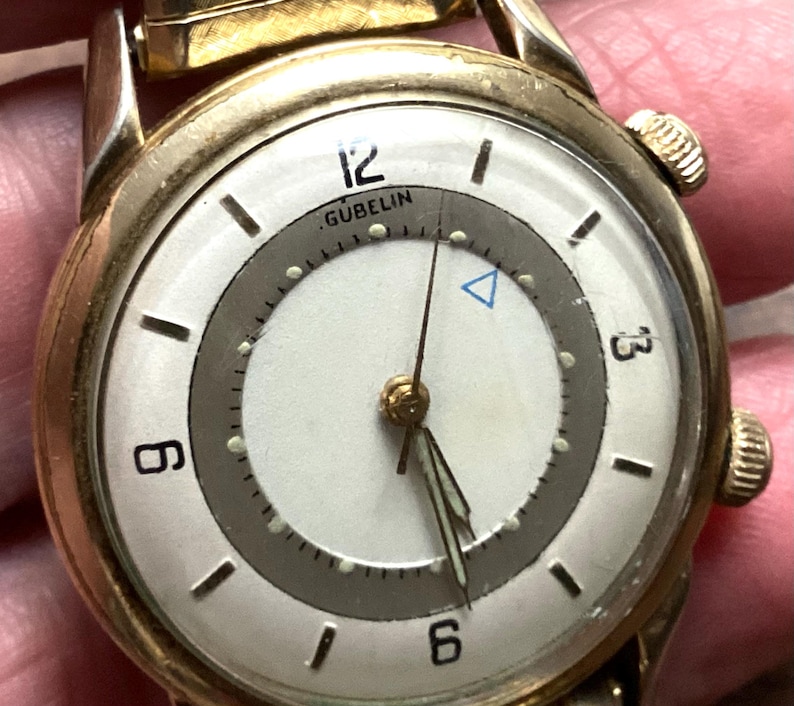 May include: A vintage gold-toned wristwatch with a white face and a gray inner ring. The watch face features black hour markers, the brand name "GUBELIN", and a blue triangle. The watch has a gold-toned band and a textured winding crown.