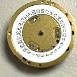 Puede incluir: Primer plano de un movimiento de reloj con un marco de metal dorado. Un anillo blanco con n&uacute;meros negros del 1 al 31 rodea el centro. El centro es dorado con peque&ntilde;os componentes mec&aacute;nicos.