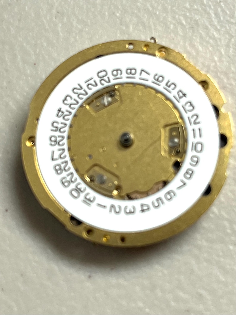 Puede incluir: Primer plano de un movimiento de reloj con un marco de metal dorado. Un anillo blanco con n&uacute;meros negros del 1 al 31 rodea el centro. El centro es dorado con peque&ntilde;os componentes mec&aacute;nicos.