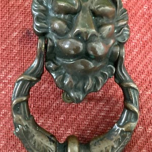 May include: Bronze lion head door knocker with a ring handle. The lion's face has detailed features, and the ring handle is decorated with a leaf and vine design. The knocker is set against a red background.