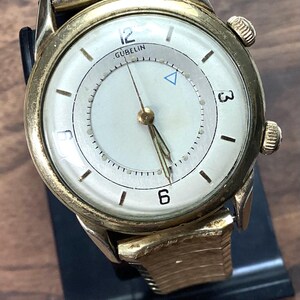 May include: Vintage gold-toned wristwatch with a white face. The watch features black numerals at 12, 3, and 6 o'clock, and a small blue triangle. The brand name "GUBELIN" is visible. The watch has a gold band and a black leather strap.