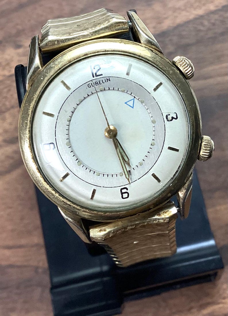 May include: Vintage gold-toned wristwatch with a white face. The watch features black numerals at 12, 3, and 6 o'clock, and a small blue triangle. The brand name "GUBELIN" is visible. The watch has a gold band and a black leather strap.