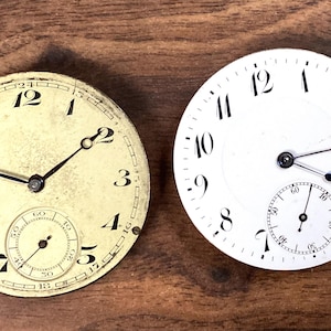 Puede incluir: Dos esferas de reloj antiguas. Una es blanquecina con números y manecillas negras, un subdial y un aspecto ligeramente envejecido. La otra es blanca con números, manecillas y un subdial negros, de apariencia más nueva. Ambas son circulares.