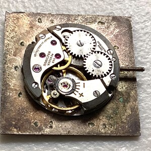 May include: Close-up of a vintage Hamilton watch movement with visible gears, jewels, and the text "Hamilton Watch Co 630". The movement is mounted on a square, textured metal base. The watch mechanism is silver and gold.