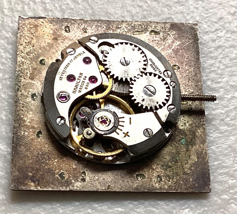 May include: Close-up of a vintage Hamilton watch movement with visible gears, jewels, and the text "Hamilton Watch Co 630". The movement is mounted on a square, textured metal base. The watch mechanism is silver and gold.