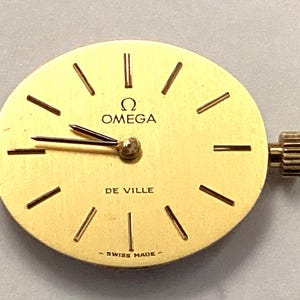 Puede incluir: Una esfera de reloj redonda de tono dorado Omega De Ville con manecillas de hora y minuto negras, marcadores de hora de bastón negros y el texto OMEGA, DE VILLE y SWISS MADE impreso en negro. Una pequeña corona dorada está en el lado derecho.