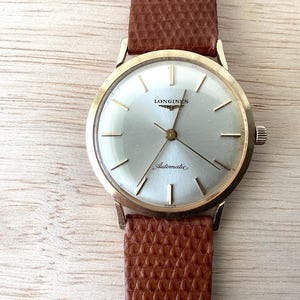 May include: A gold-tone Longines automatic wristwatch featuring a round silver dial with thin gold-tone hour markers and hands. The watch has a textured brown leather strap with a distinct grain pattern and a small crown on the right side.