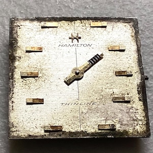 May include: A vintage Hamilton watch face with a silver-toned square dial. The face features gold-toned hour markers, hands, and the brand name "HAMILTON". The word "THINLINE" is printed below the center. The watch face shows signs of age.