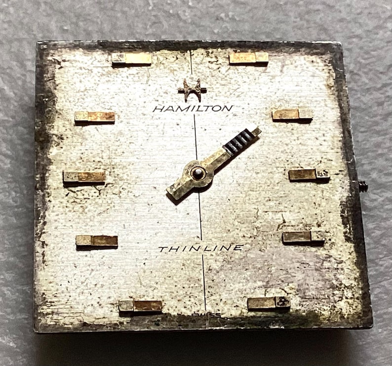 May include: A vintage Hamilton watch face with a silver-toned square dial. The face features gold-toned hour markers, hands, and the brand name "HAMILTON". The word "THINLINE" is printed below the center. The watch face shows signs of age.