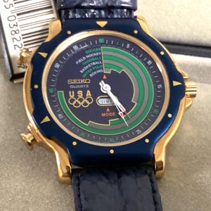 May include: A Seiko Quartz watch with a gold-tone case, navy blue bezel, and a navy blue leather strap. The watch face features the Olympic rings, the text "USA", and a multi-colored dial with sports indicators.