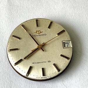 May include: A round, off-white Movado Kingmatic S watch dial with gold-tone hour markers and hands. The dial features a date window at the three o'clock position displaying the number 17, the Movado logo, and the text T SWISS T at the bottom.