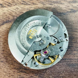 May include: Close-up of an Elgin watch movement with intricate gears, wheels, and a ruby jewel. The silver-toned mechanism is partially exposed, revealing the inner workings of the watch. The text "ELGIN" and "SWISS" are visible.