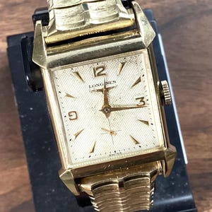 Puede incluir: Un reloj Longines vintage dorado con esfera rectangular. El reloj presenta una esfera color crema con manecillas e indicadores dorados, incluidos los números 12, 3 y 9. La correa del reloj también es dorada y tiene un diseño texturizado único.