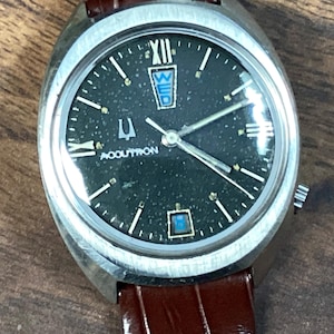 May include: A vintage Bulova Accutron watch with a dark speckled dial, silver case, and brown leather strap. The watch face displays the day of the week and the Accutron logo. The watch has a silver bezel and a brown leather strap.