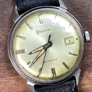 Op de afbeelding: Een vintage Bulova polshorloge met een zilverkleurige kast en een crèmekleurige wijzerplaat. Het horloge heeft zwarte wijzers met lichtgevende accenten, een datumweergave op 3 uur en een zwarte leren band. Het woord "WATERPROOF" staat op de wijzerplaat.
