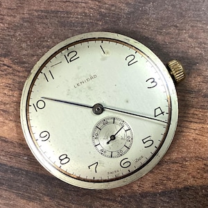May include: A vintage watch face with a cream-colored dial, black numerals, and a small seconds dial. The watch face has a gold-colored bezel and a winding crown. The text "LENIDAD" is printed on the face. The watch is marked "SWISS MADE".