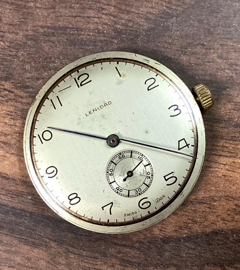 May include: A vintage watch face with a cream-colored dial, black numerals, and a small seconds dial. The watch face has a gold-colored bezel and a winding crown. The text "LENIDAD" is printed on the face. The watch is marked "SWISS MADE".