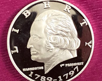 May include: A silver-colored coin featuring a profile of George Washington. The coin's edge has a textured design. The words "LIBERTY" and "WASHINGTON" are visible, along with the dates "1789-1797" and "1st PRESIDENT".