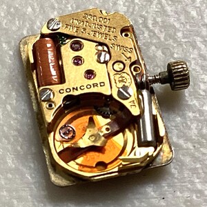 May include: Close-up of a gold-toned watch movement with the text "CONCORD" and "SWISS". The movement has small gears, jewels, and a winding stem. The text "280.001 UNADJUSTED FIVE 5 JEWELS" is also visible.