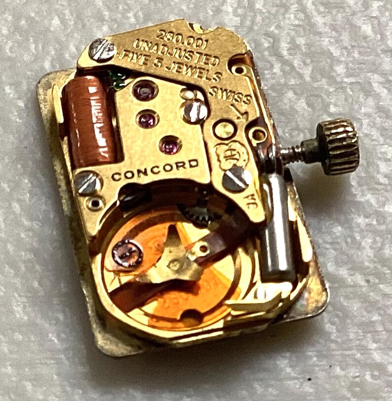 May include: Close-up of a gold-toned watch movement with the text "CONCORD" and "SWISS". The movement has small gears, jewels, and a winding stem. The text "280.001 UNADJUSTED FIVE 5 JEWELS" is also visible.