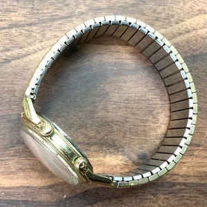 May include: A gold-toned wristwatch with a silver-colored, expandable band. The watch face is a light color, and the watch has a small crown. The band is made of small, rectangular metal pieces. The watch is on a wooden surface.