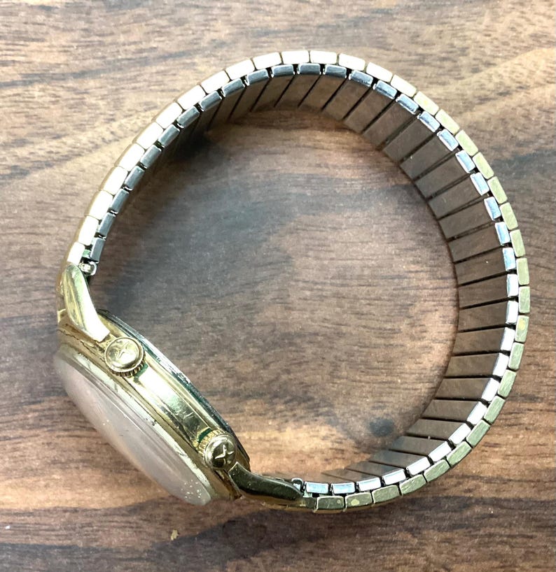 May include: A gold-toned wristwatch with a silver-colored, expandable band. The watch face is a light color, and the watch has a small crown. The band is made of small, rectangular metal pieces. The watch is on a wooden surface.