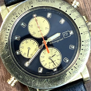 May include: A gold-toned quartz chronograph watch with a black face and three sub-dials. The watch features a tachymeter bezel, date display, and a black leather strap. The watch hands are orange and gold.