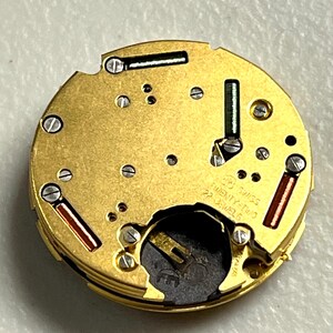 Puede incluir: Primer plano de un movimiento de reloj dorado con detalles intrincados. Presenta peque&ntilde;os tornillos, acentos verdes y rojos, y el texto "SWISS TWENTY-TWO 22 JEWELS". El mecanismo circular revela una compleja estructura interna.