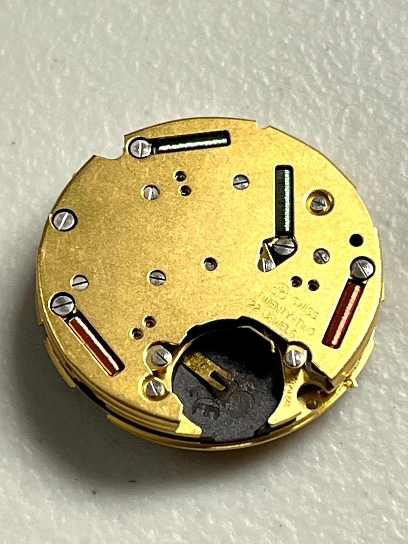 Puede incluir: Primer plano de un movimiento de reloj dorado con detalles intrincados. Presenta peque&ntilde;os tornillos, acentos verdes y rojos, y el texto "SWISS TWENTY-TWO 22 JEWELS". El mecanismo circular revela una compleja estructura interna.