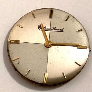 Puede incluir: Una esfera de reloj redonda Lucien Piccard con un acabado bicolor en plata y champán. La cara incluye marcadores de hora en tono dorado y manecillas de hora y minutos a juego en tono dorado. El nombre de la marca Lucien Piccard está impreso en negro.