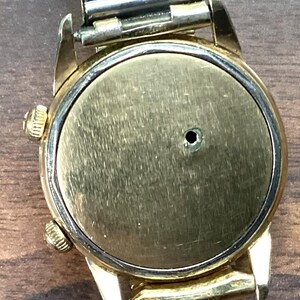 May include: A vintage gold-toned wristwatch with a silver-colored metal band. The watch face is a brushed gold color with a small hole in the center. The watch has two small gold-toned knobs on the side.