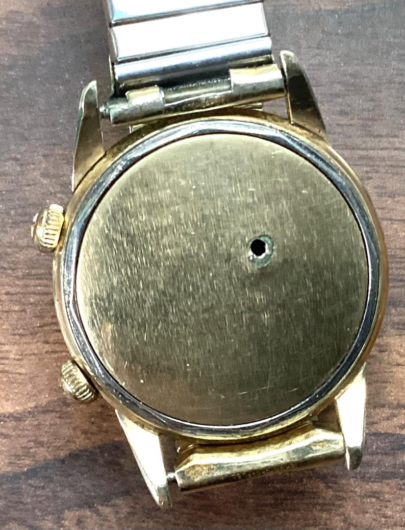 May include: A vintage gold-toned wristwatch with a silver-colored metal band. The watch face is a brushed gold color with a small hole in the center. The watch has two small gold-toned knobs on the side.