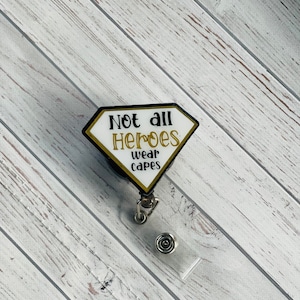 May include: A black and white retractable badge holder with a gold diamond-shaped charm that reads "Not all heroes wear capes".