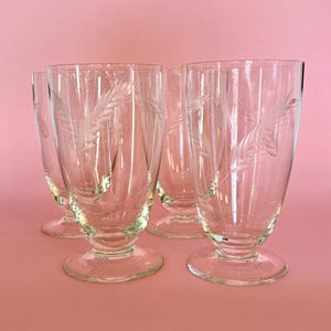 May include: A set of four clear glass tumblers with a footed base, each featuring a delicate etched wheat stalk pattern on the exterior surface. The glasses have a smooth, cylindrical body that tapers slightly toward the base.