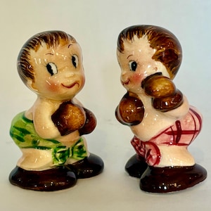 Puede incluir: Dos figuras de boxeadores de cerámica vintage. Cada figura es un niño sonriente con cabello castaño, mejillas rosadas y guantes de boxeo marrones. Uno lleva calzoncillos verdes, el otro calzoncillos a cuadros rojos. Las figuras tienen bases de color marrón oscuro.